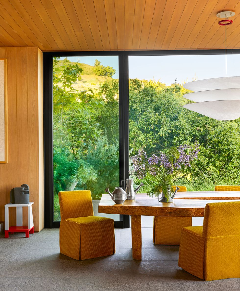 a dining room has wood walls and sliding glass doors overlooking a verdant hillside, a bleached hardwood table with armless chairs in a mustard yellow fabric, a three layered suspension light, and a stone floor