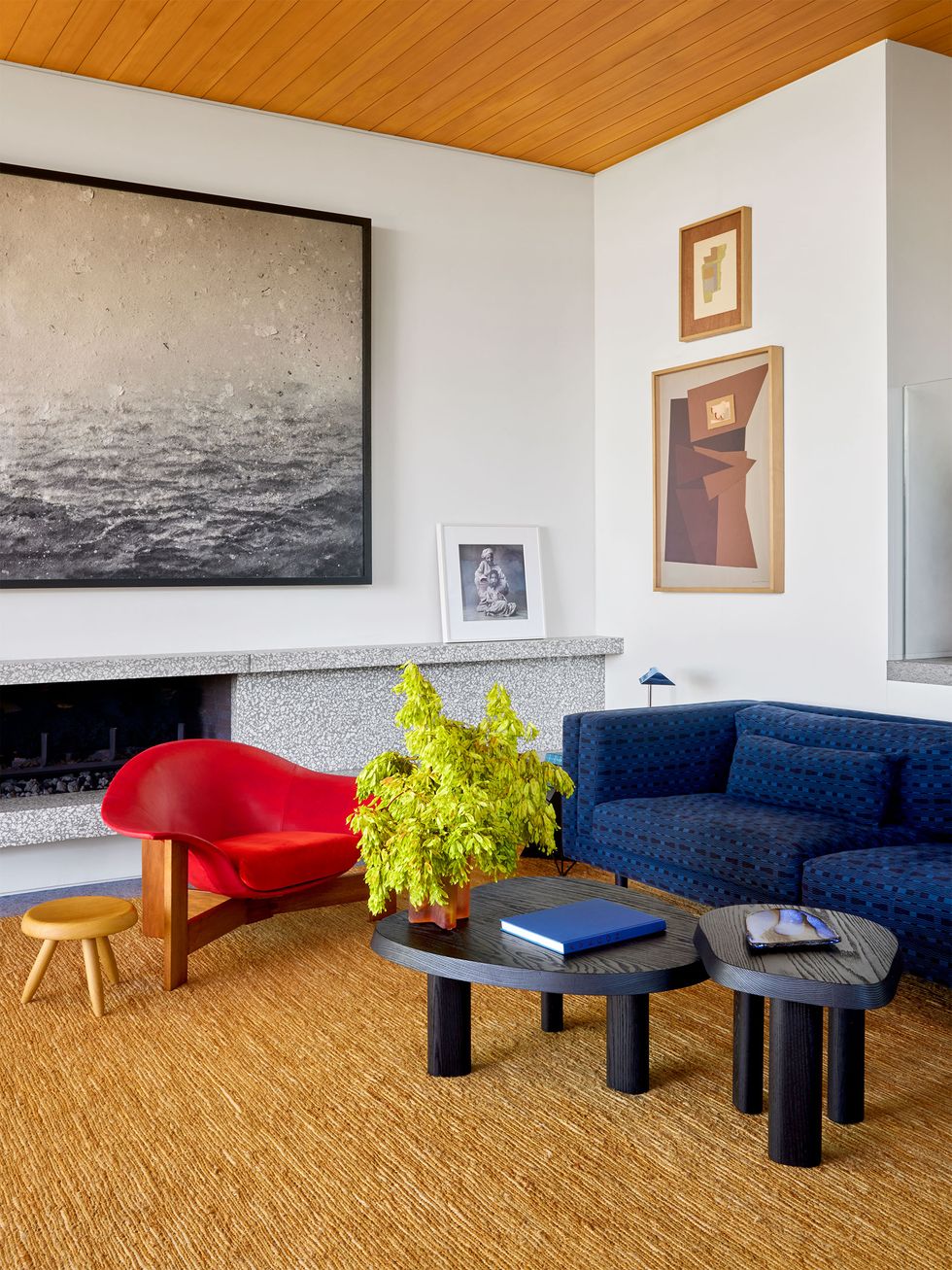 a living room has white walls, a low gray stone fireplace, a jute rug, two three legged cocktail tables of different heights, a modern red armchair with a wood base, a deep blue sofa, and multiple artworks