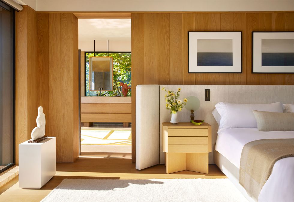 a bedroom with wood floors and walls has a light upholstered bed with headboard that wraps around a nightstand, framed photos above bed, a white rug, and a small white side table with a white sculpture