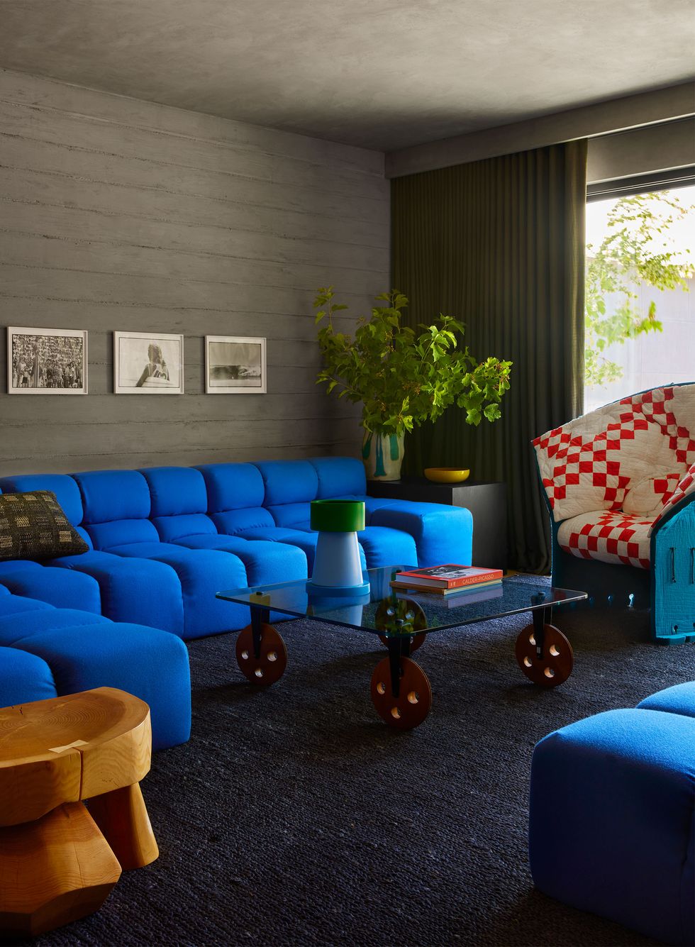 media room has a faux concrete wall and a curtained sliding glass door, bright blue sectionals, a glass table with buttonlike legs, a wood side table, a teal chair with red and white checked fabric, charcoal rug
