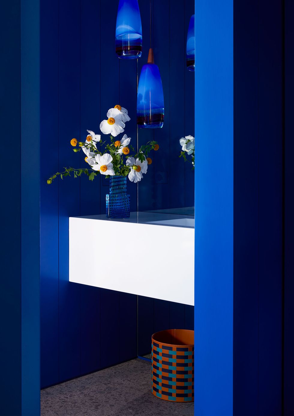 a powder room is painted in a deep but bright blue, a blue vase with white and orange flowers sits on a white sink, three glass oblong pendants hang over the sink, a pop art basket sits under the sink