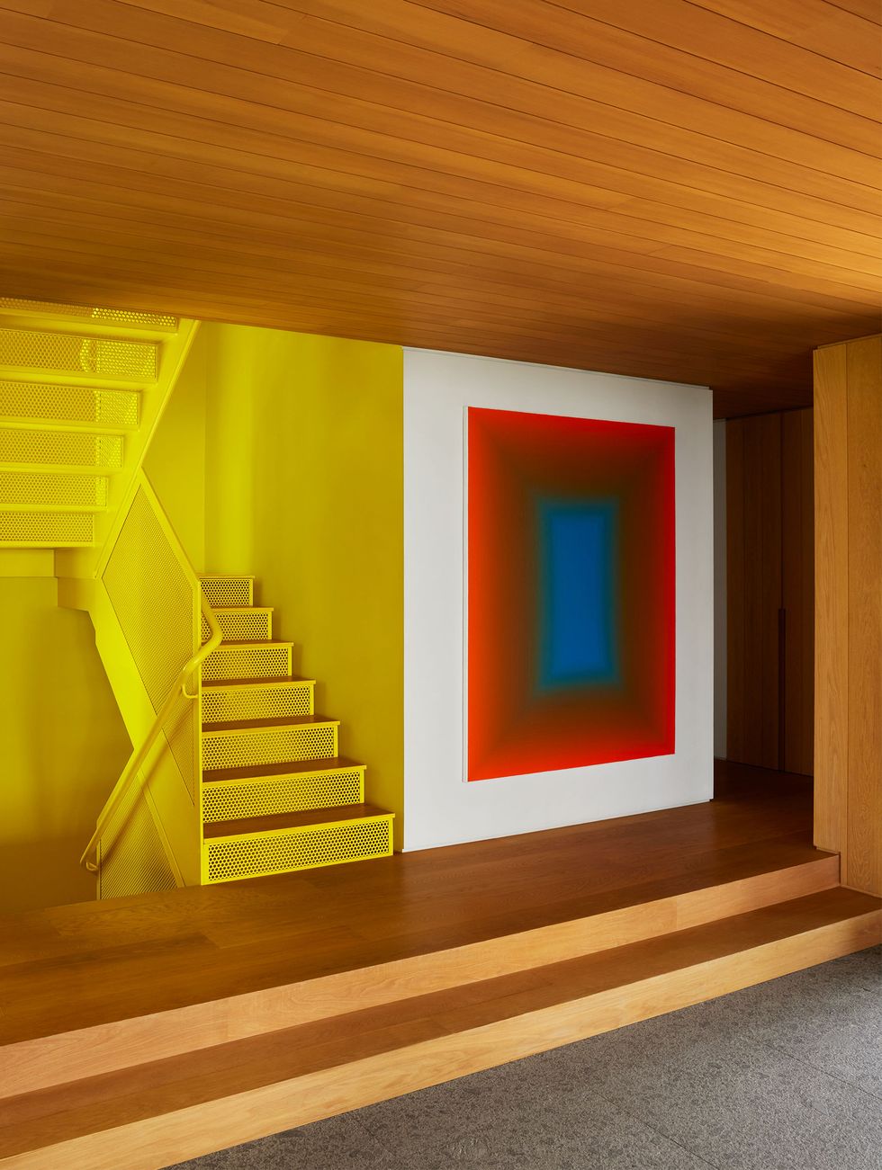 a foyer has two steps up to bright yellow industrial style staircase, wood floor, walls, and ceiling, and a large rectangular painting with a wide red border and a narrow green border around a blue center