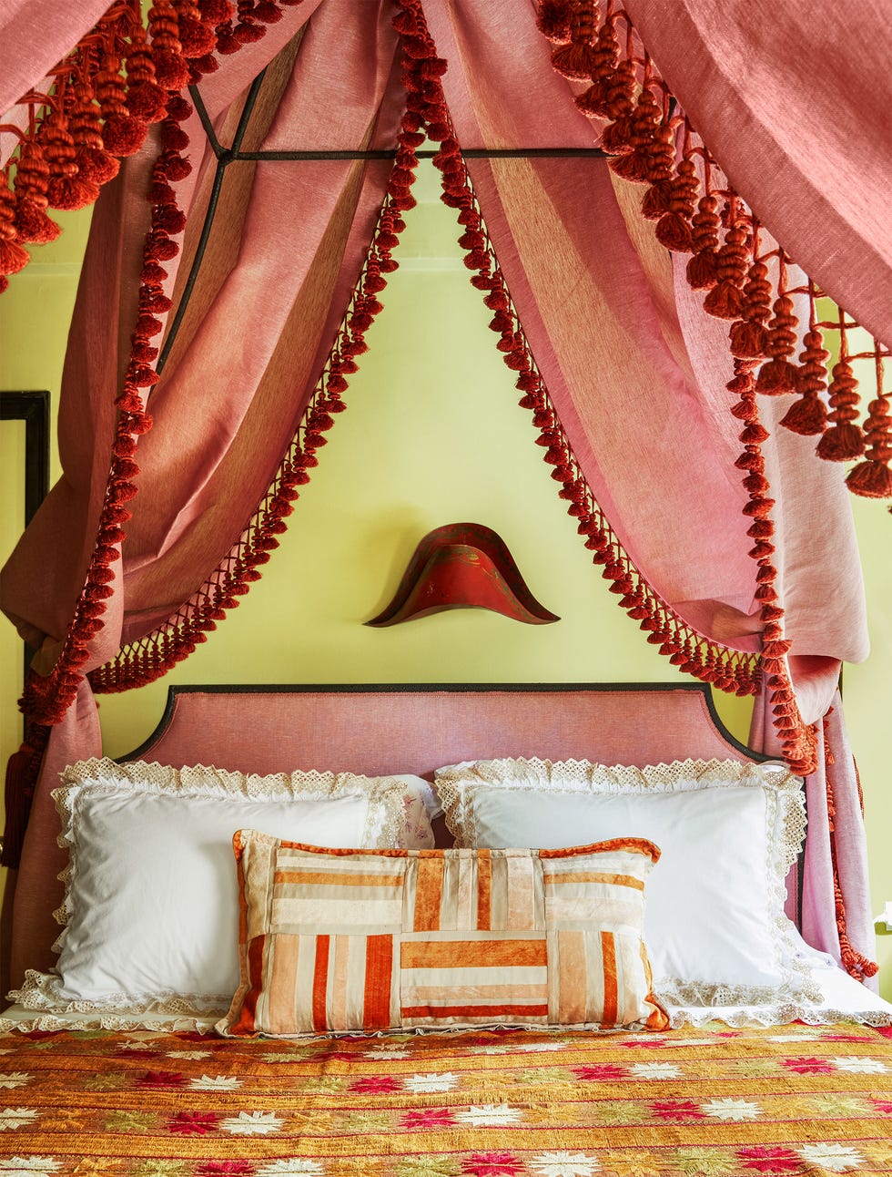 le lit avec une tête de lit et un baldaquin assortis en tissu rose avec une bordure à pompons tout autour, un couvre-lit à motifs de couleur safran, des oreillers bordés de dentelle et une applique au-dessus de la tête de lit