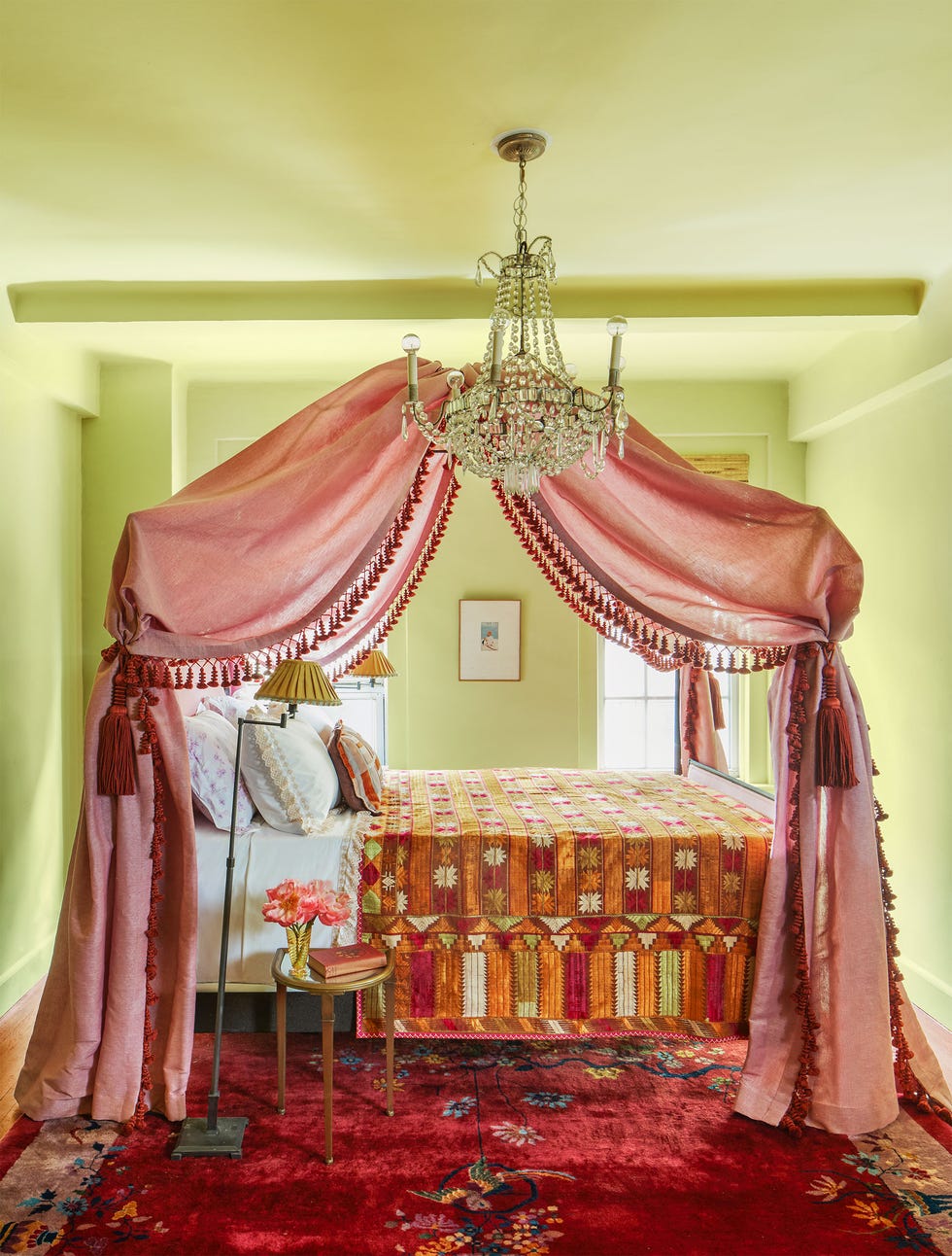 une petite pièce aux murs de sauge comprend un lit près de deux fenêtres avec un baldaquin en tissu perlé de couleur rose et une couverture multicolore, deux lampadaires, une table de chevet à trois pieds et un lustre en cristal