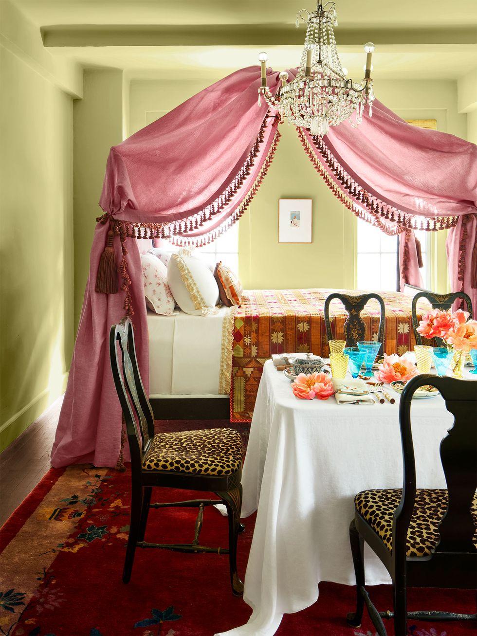a small room with sage walls has a bed near two windows with a beaded, rose colored fabric canopy and a multicolored spread, a crystal chandelier, and a set dining table and chairs with leopard seats