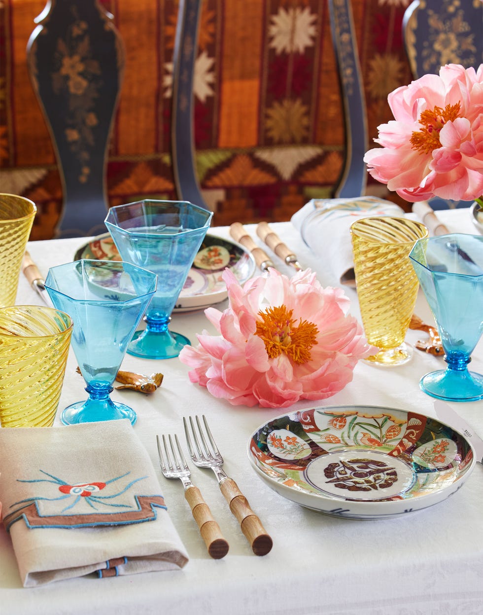 in a closeup of a table covered in a white cloth are patterned shallow bowls, forks with bamboo handles, cloth napkins, amber drinking glasses with a spiral design and hexagonal shaped aqua glasses