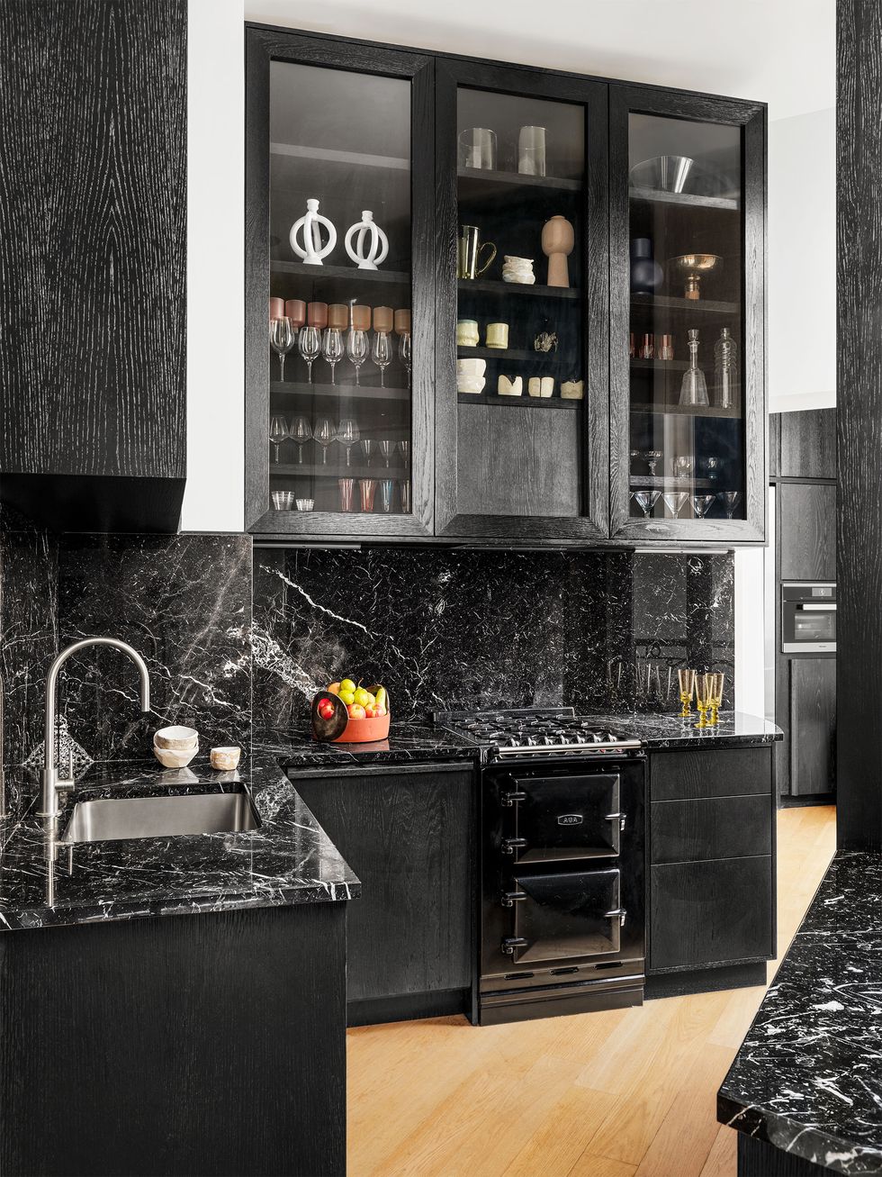 a kitchen has black lower cabinets topped with white veined black marble and upper cabinets with glass fronts showing glassware and dishes, stainless steel sink, and planked oak flooring