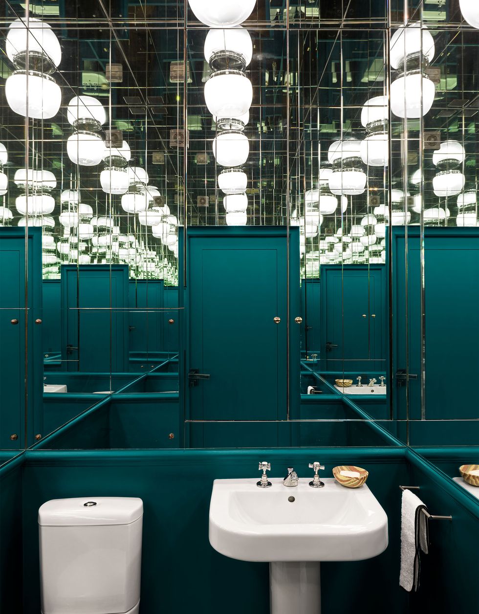 a bathroom has white porcelain fixtures and walls that are painted a deep teal, a mirror on one wall and mirrors on the ceiling reflect multiple images of the room and of the vintage opal glass ceiling light