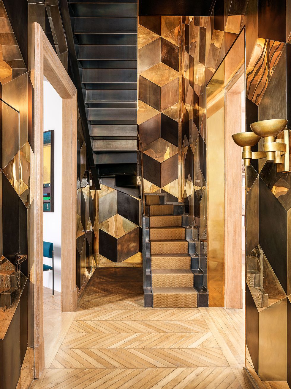 un hall d'escalier a des murs recouverts de plaques de laiton polies et patinées selon un motif de cubes imbriqués, deux appliques en laiton, un plancher en bois à motif de chevrons