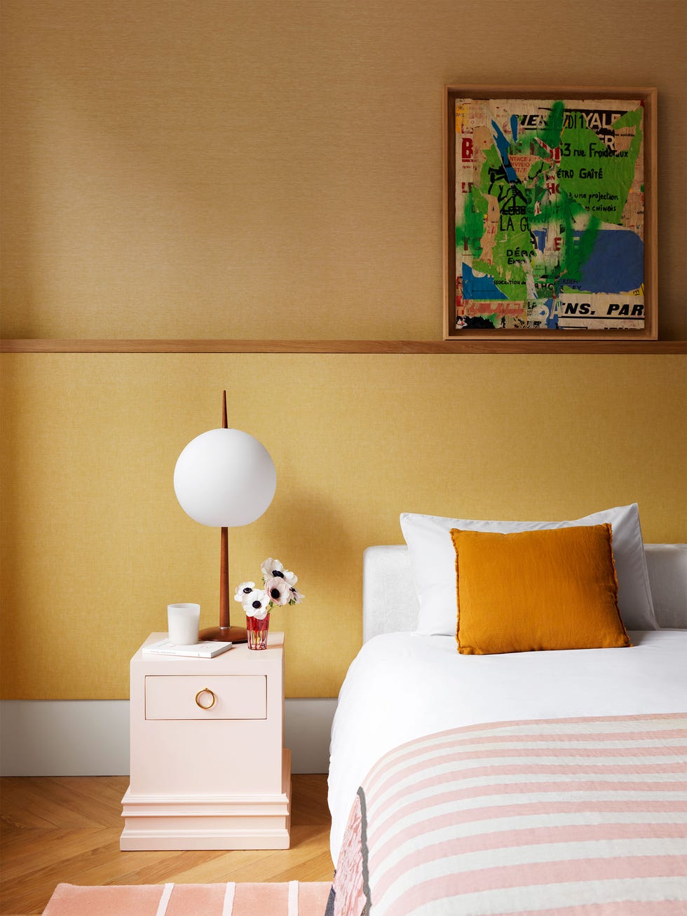 a bedroom has mustard color walls with a collagelike artwork above the bed, white headboard and linens, pink and white striped bedcover, pink nightstand with a globe lamp, small pink and white striped rug