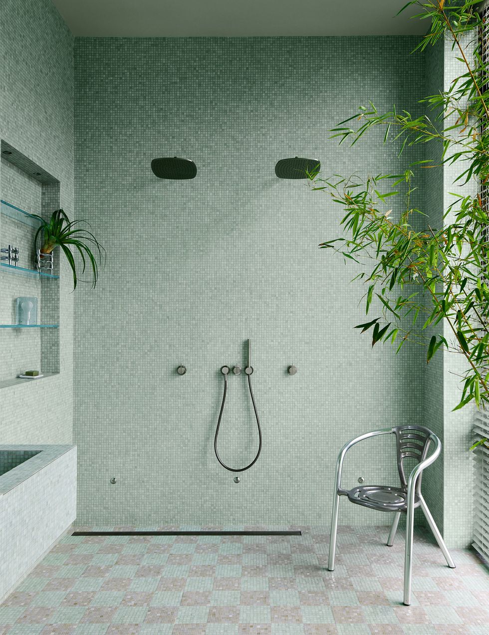 the walls of a bathroom are done in very small sea foam colored square glass tiles and the floor has larger two toned squares, inset glass shelves, dual rain showerheads, and a vintage aluminum chair