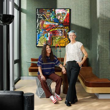 sue bird sits on a black and white patterned stool with megan rapinoe standing beside her in a dining room with a glass table and brown suede banquette against green textured walls and a colorful abstract painting