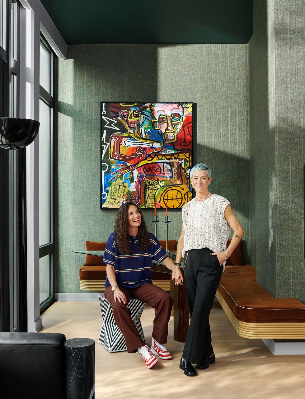 sue bird sits on a black and white patterned stool with megan rapinoe standing beside her in a dining room with a glass table and brown suede banquette against green textured walls and a colorful abstract painting