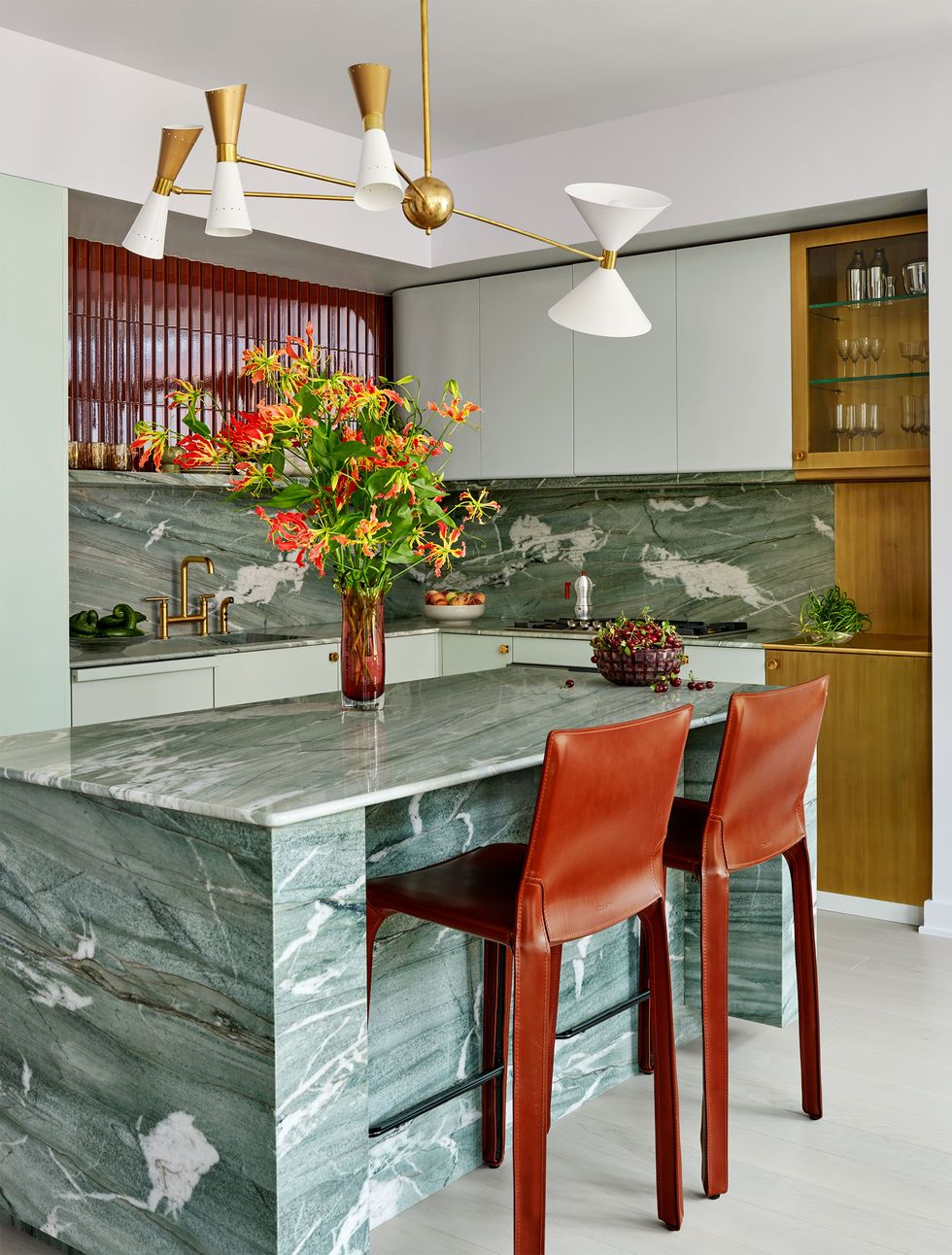 in a kitchen is a light green marble island with two brownish red leather barstools and a four armed chandelier with ivory gold shades, marble backsplash and shiny brown tiles above and light green cabinets