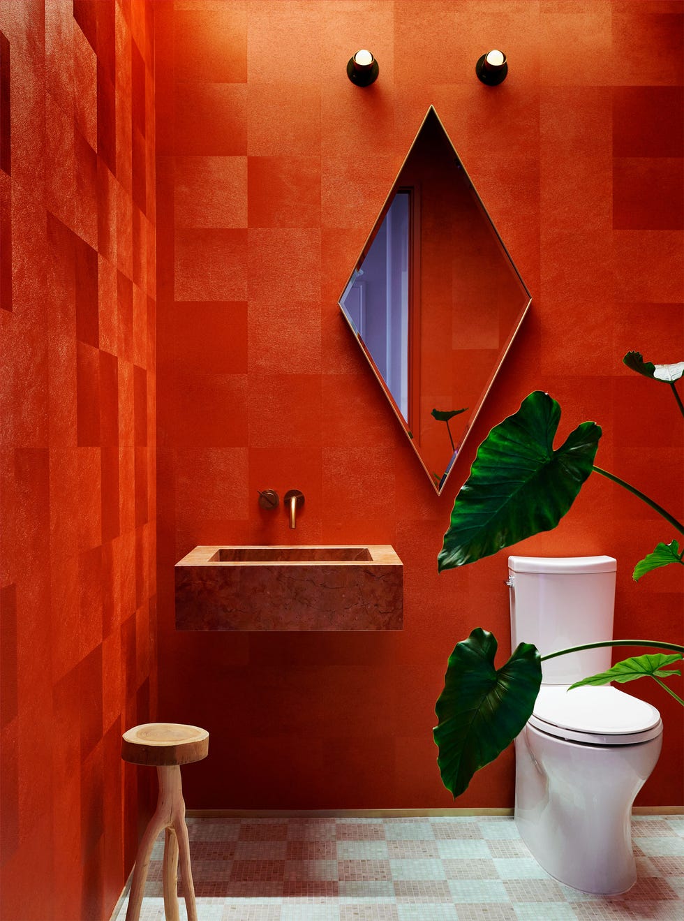 the walls of a powder room are covered in a textured red wallpaper, a square free hanging sink is in red travertine, diamond shaped mirror with two small sconces above, a small three legged wooden stool