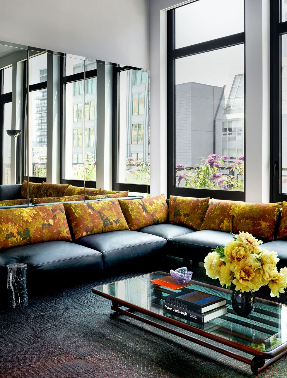 a living room framed by mirrors and windows has a sectional with black leather seat cushions and flowered fabric back cushions, a low glass cocktail framed in curved wood, and a dark patterned rug