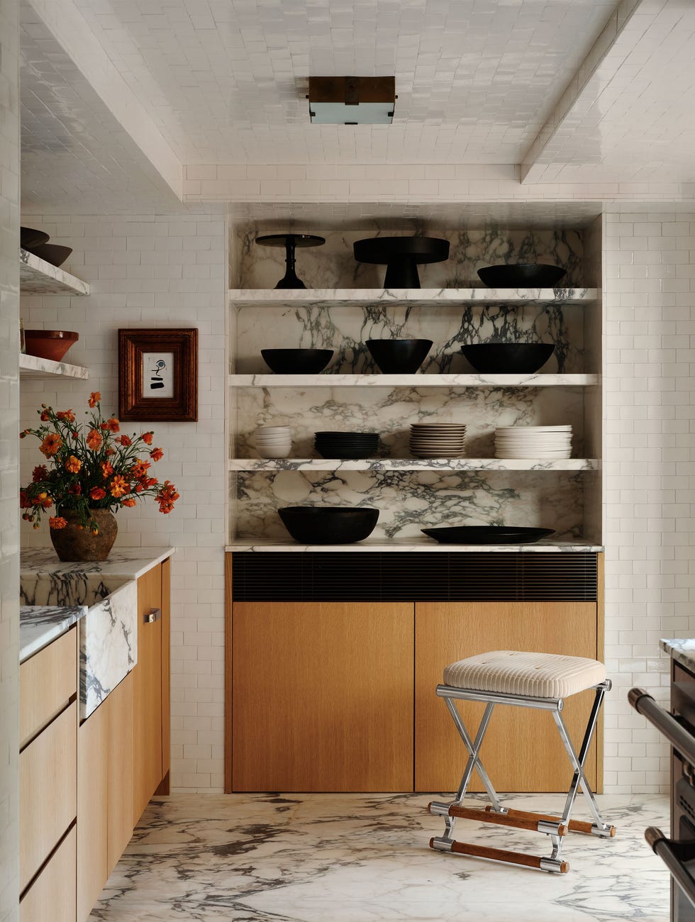 Kitchen walls feature white subway tile, light marble recessed open shelves with dark veining to showcase black and white tableware, light wood cabinets with marble sink and countertops, marble flooring, square ceiling lights