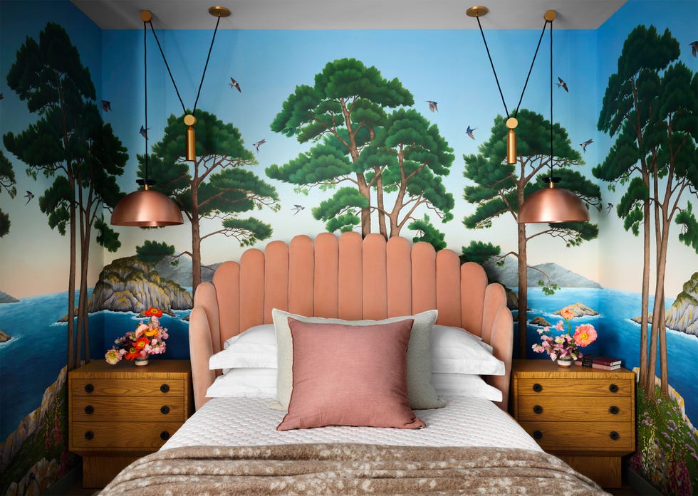 a bed with a curved headboard upholstered in a light salmon colored fabric, wooden nightstands with drawers, two copper hooded pendants, wallpaper in a landscape of birds, evergreens, rocks, and blue sea