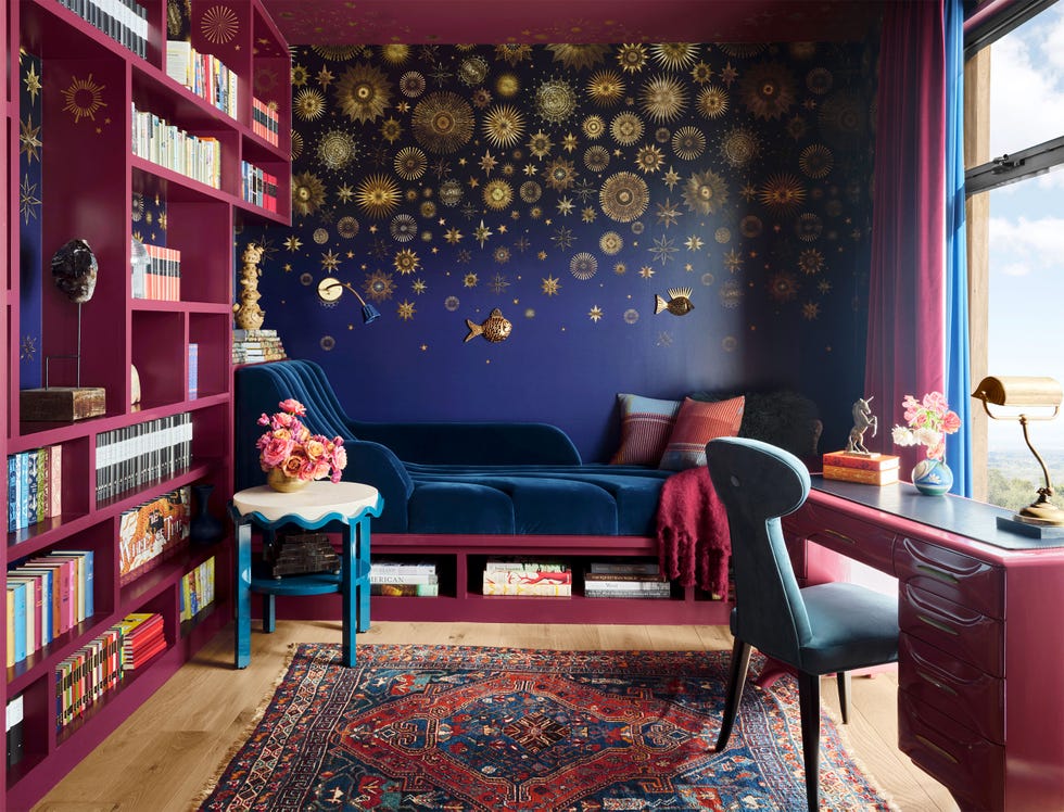 a study with magenta colored bookshelves, desk, base of daybed, and curtains, deep blue velvet on daybed, blue wall with gold sun designs and 3d fish, turquoise side table with white top, light blue desk chair, colorful rug