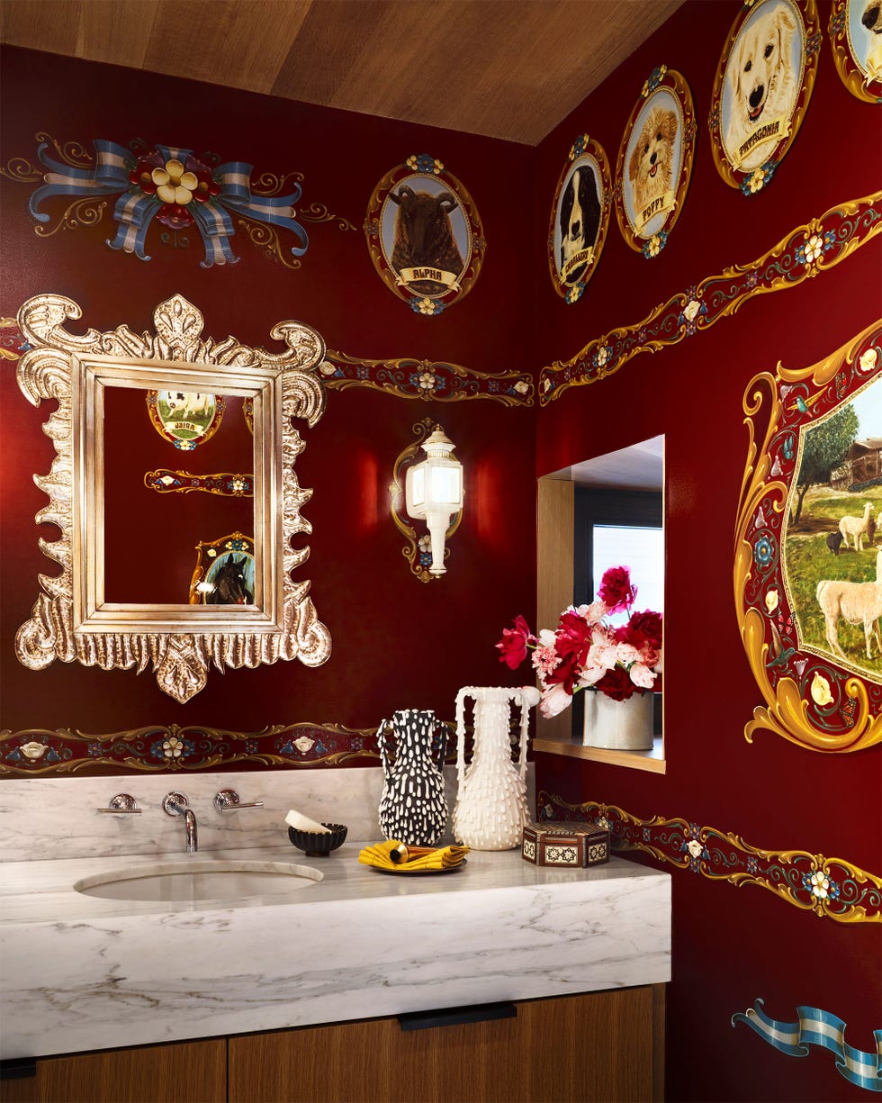 a powder room has dark burgundy walls adorned with framed pictures of animals, decorative mirrors, and ribbons, and trim, white marble sink and counter with vases, soap and towels, silver fittings