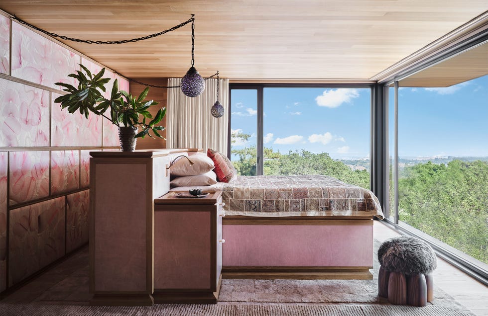 a bedroom with two glass walls, an upholstered wall, bed with built in dresser and side tables, pink base, muted print bedcover, small side sconces and dangling globe pendants, fur topped wood stool, wood ceiling, rug