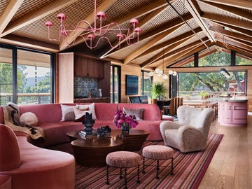 living room with slanted, beamed wooden ceiling, curved pink sectional sofa, curved cocktail table, boucle chair, two stools, striped rug, pink chandelier, large windows, dining table and kitchen island in background