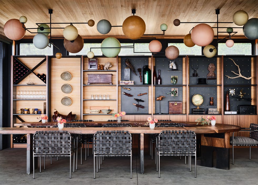 dining area with u shaped wood table and webbed chairs with fabric seats, niches on wood wall filled with tools, art objects, glasses and wines, three chandeliers with globes of different sizes and colors