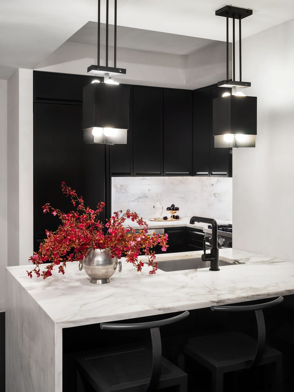 a small kitchen with a white marble backsplash and counter with a sink and black fittings  silver vase with red flowers  two