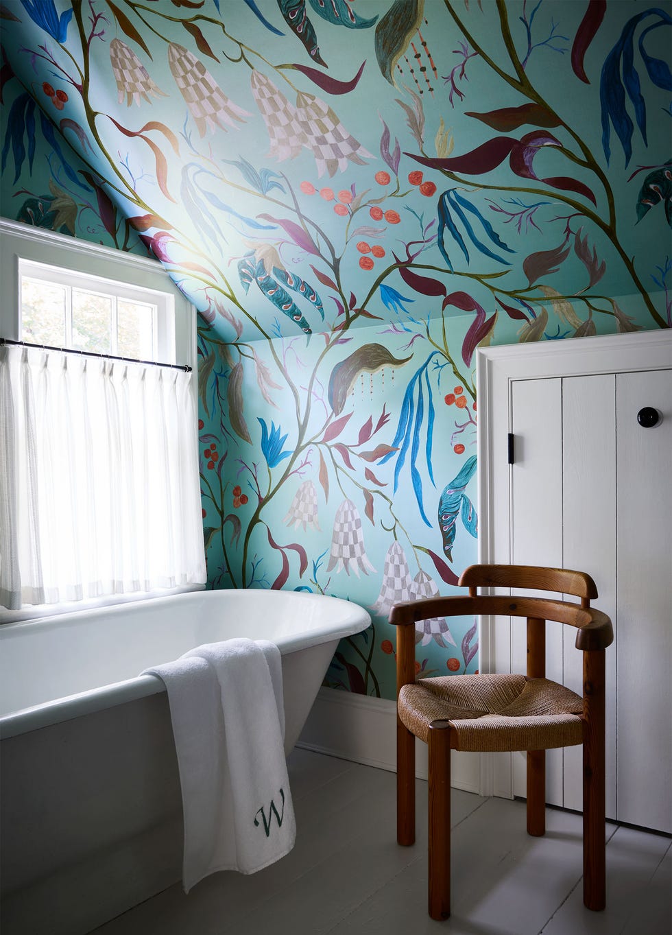 Bathroom with a deep white bathtub and a white towel, a wooden chair with a curved back and a rush seat, a whimsical wallpaper with an aquamarine background and colorful leaves, vines and sea creatures, a window with a white curtain