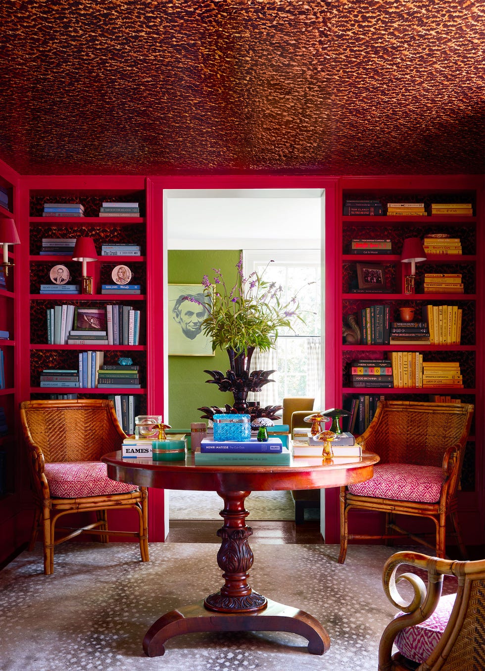 a library with magenta bookshelves full of books and small pictures, wall lamps with shades attached to the shelves, a round wooden table with three rattan chairs with printed cushions, a wallpapered ceiling