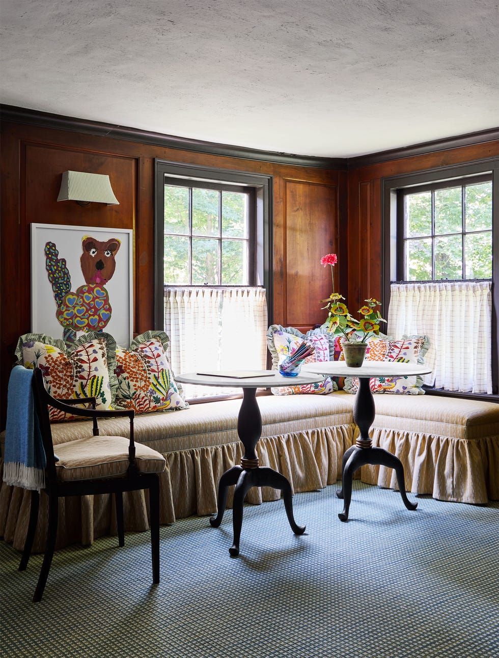 Family room with wood paneled walls, two windows with half curtains below, built-in bench with skirt and colorful accent pillows, one chair and two round tripod tables, framed artwork of a child on the wall