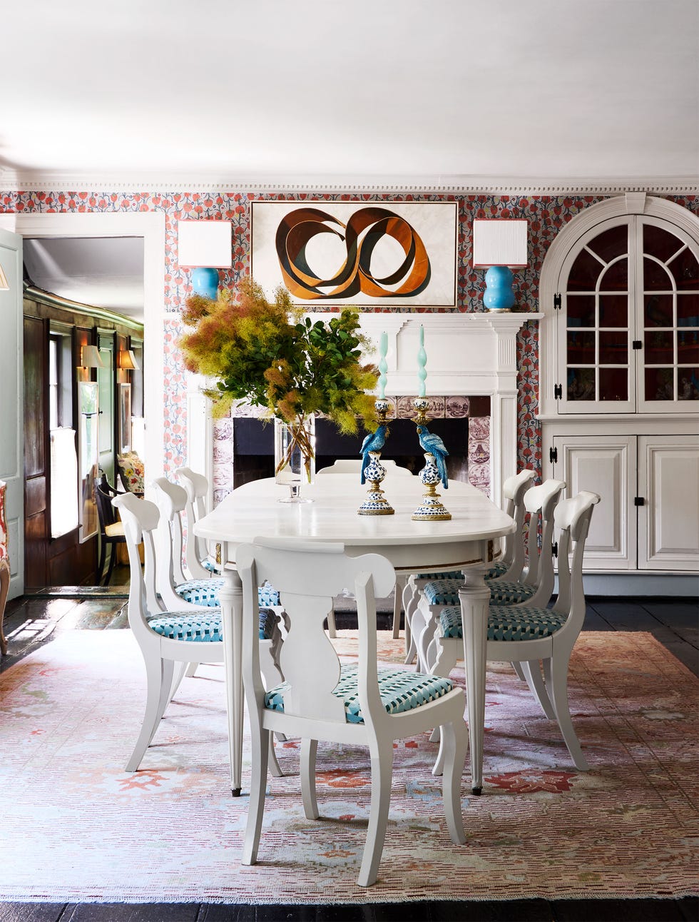 a dining room with a white oblong table and chairs with turquoise patterned fabric seats, fireplace with decorative tile inlays and white mantel with two lamps on it and a piece of art above, built-in arched cabinet