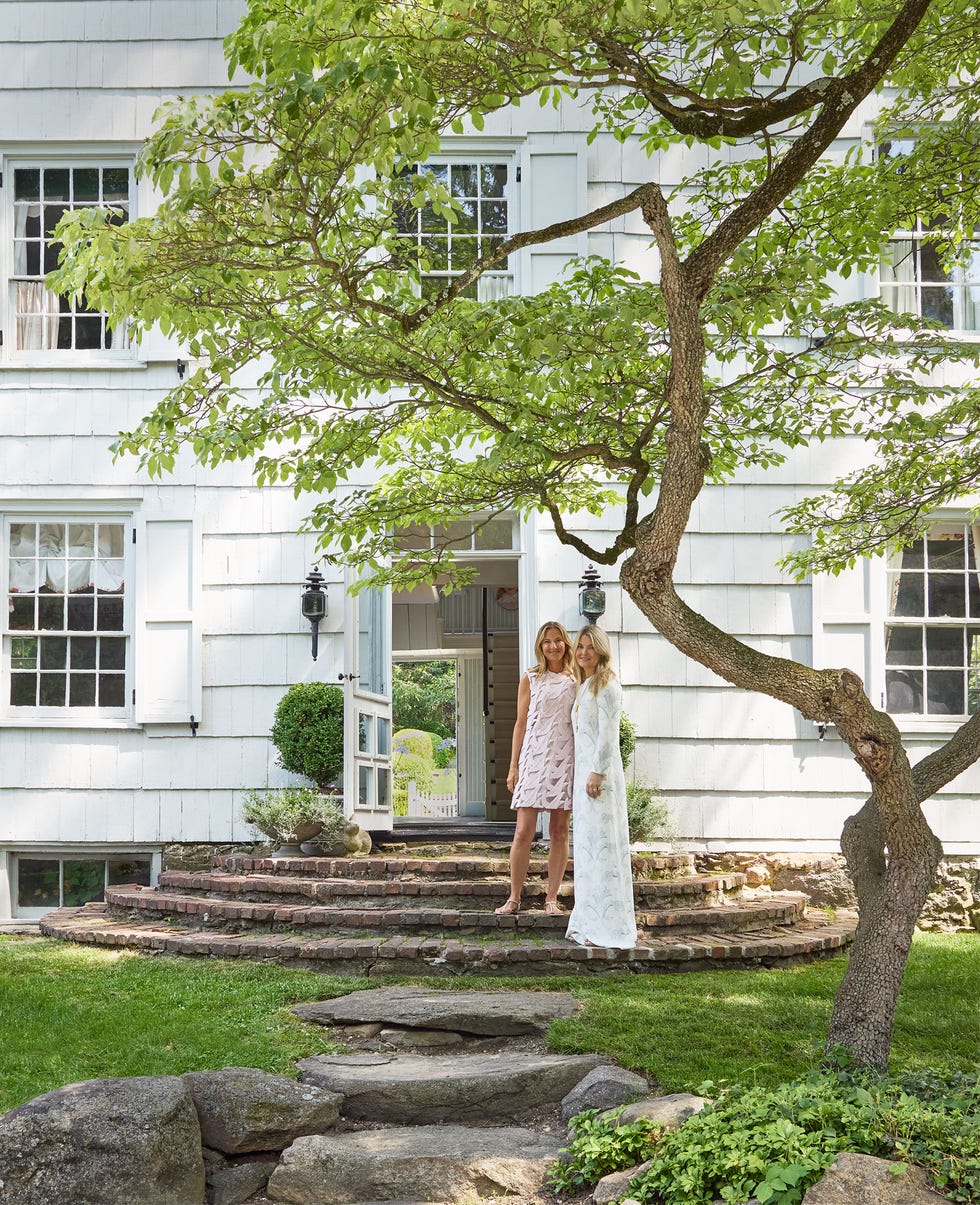 In front of a white clapboard house with semicircular brick steps leading up to an open door stand two women, both with long blonde hair, one in a short pink dress and the other in a long white dress, rocks and a tree in the yard