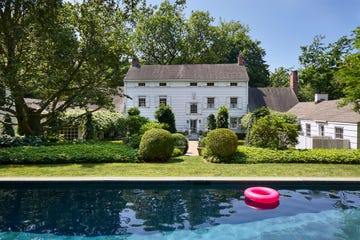 the exterior of a 17th century three story white clapboard house with two wings and an out building surrounded by trees and landscaped shrubs, pool in foreground with a pink floating tube