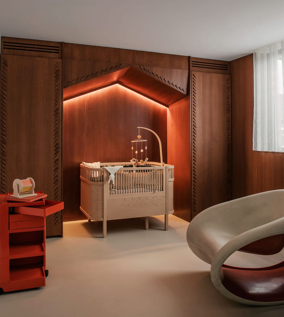 a wood paneled nursery has embellishments around a niche that houses a light wood crib with mobile attached and subtle interior lighting, red trolley with drawers, an armless molded taupe rocking chair