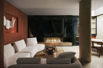 a living room with a wood paneled wall with a white 3d artwork, curved white sofa and a taupe colored loveseat, glass and stone cocktail table, supporting column, black zinc wall with fireplace, stone hearth