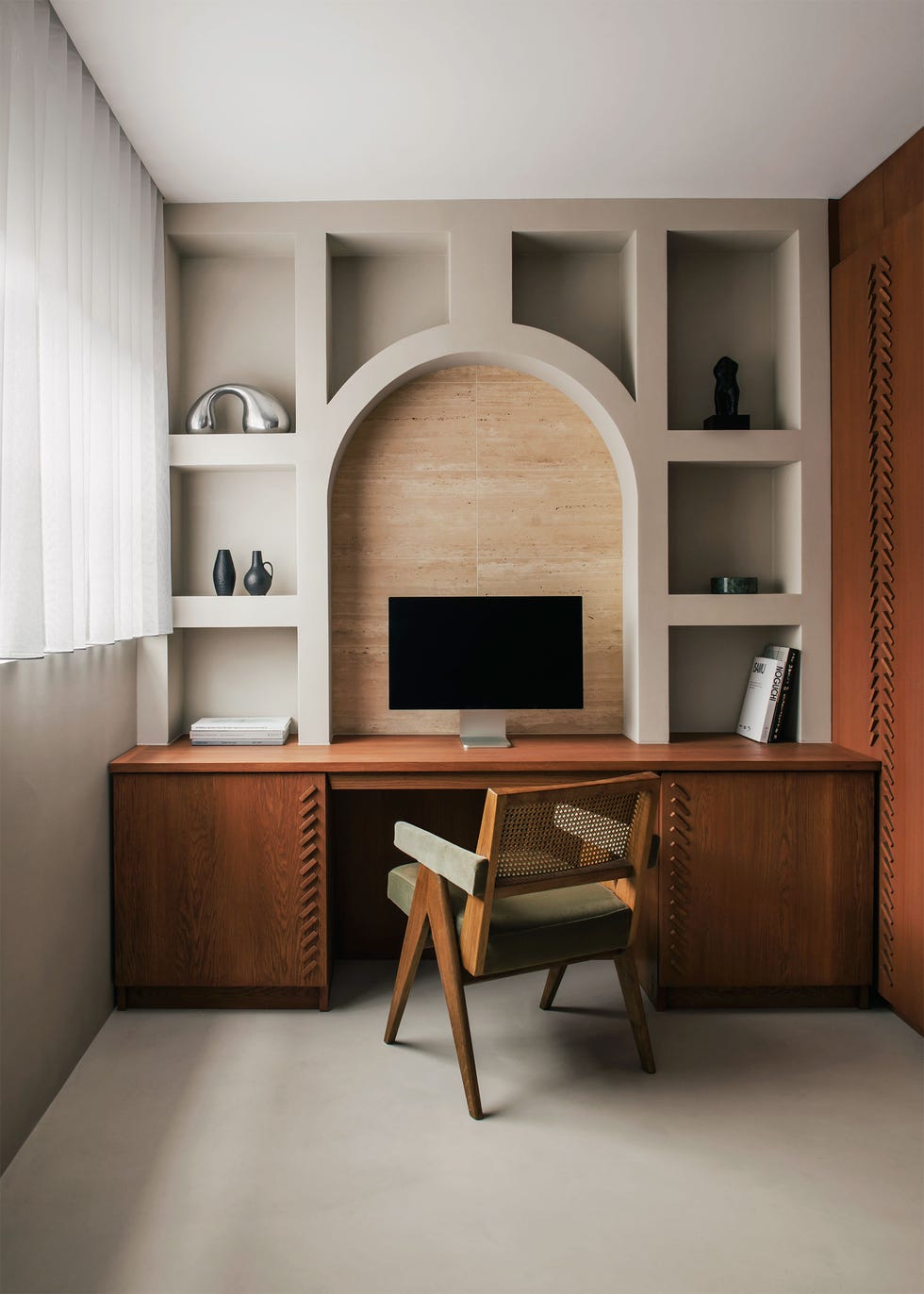 a built in desk in the same wood as wall, a computer monitor in an arched niche with cubbylike shelves around it holding art objects, a pierre jeaneret chair with olive cushion, taupe rug, white curtained window