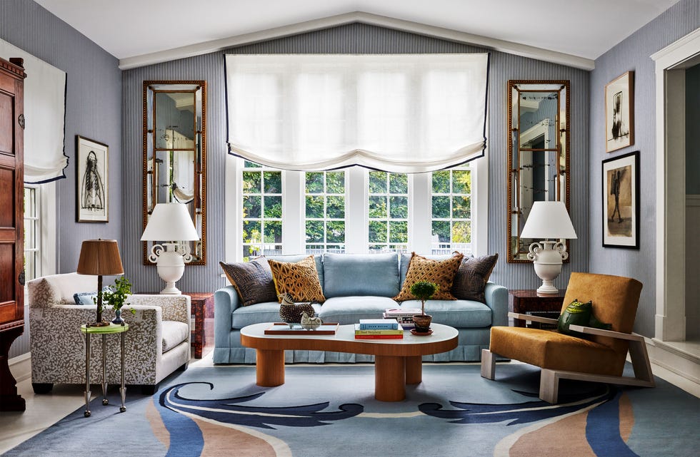 blue sofa with accent pillows flanked by end tables with white lamps, two armchairs and a side table, cocktail table, large paned window with white roman shade, matching mirrors on each side, blue rug