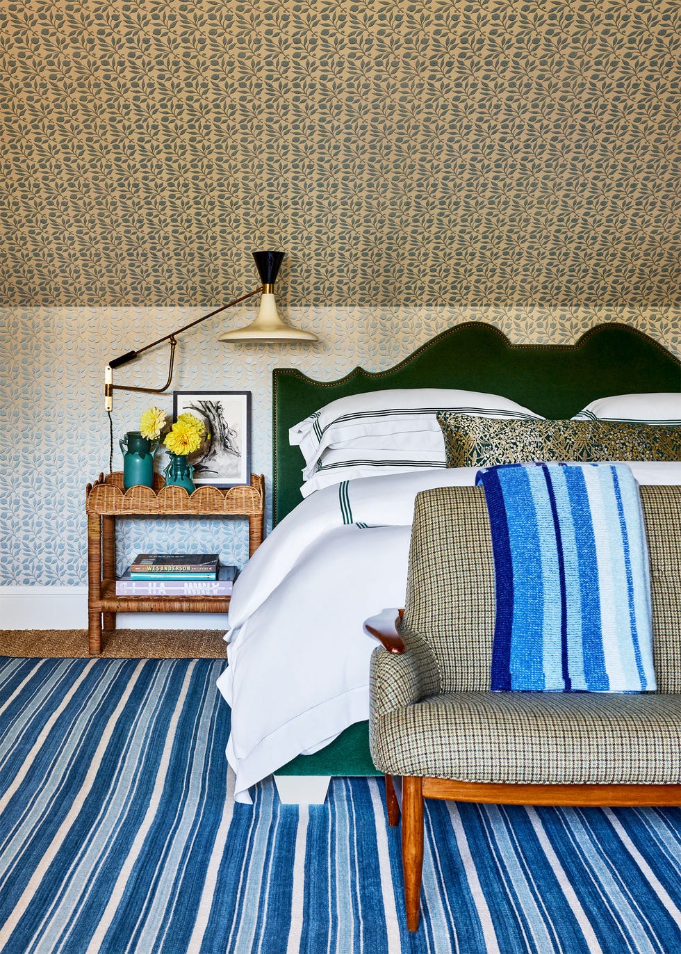 bedroom with walls and slanted ceiling covered in a small blue pattern, emerald colored fabric headboard, blue and white striped rug, check pattern on settee at foot of bed, rattan nightstand with lamp