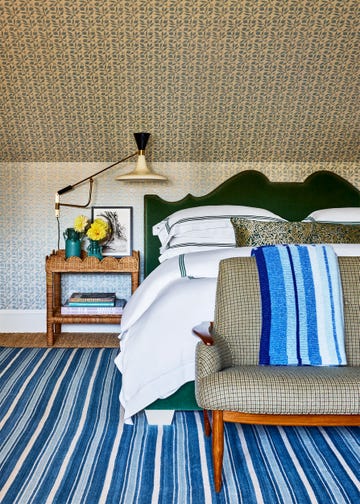 bedroom with walls and slanted ceiling covered in a small blue pattern, emerald colored fabric headboard, blue and white striped rug, check pattern on settee at foot of bed, rattan nightstand with lamp