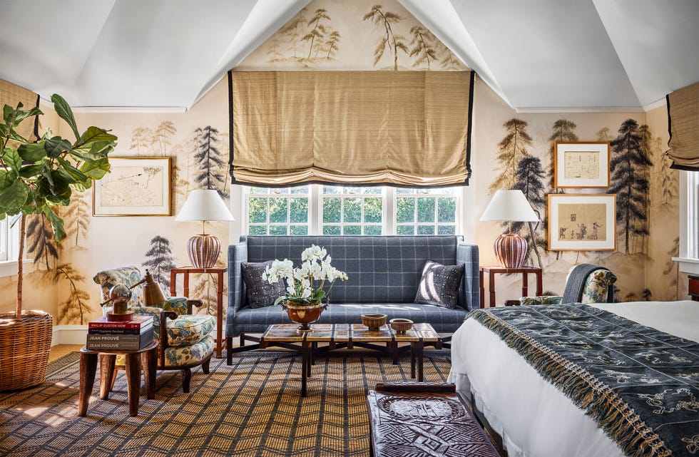 tree patterned wallpaper in a bedroom, sofa in gray and white square fabric in front of window with beige roman shade, two chairs in flower fabric, end tables with lamps, cocktail table, bed with patterned blanket