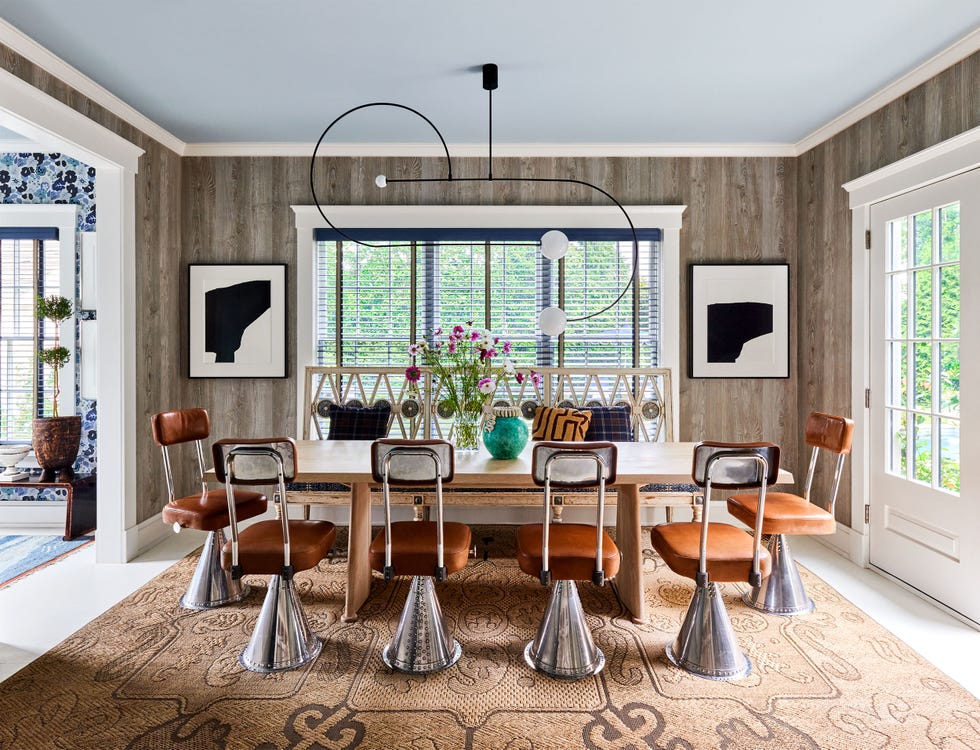 dining area with rectangular table and six chairs plus a bench on one side, gray paneled walls, wrought iron and globe chandelier, large windows on two sides, black and white artworks, natural patterned rug