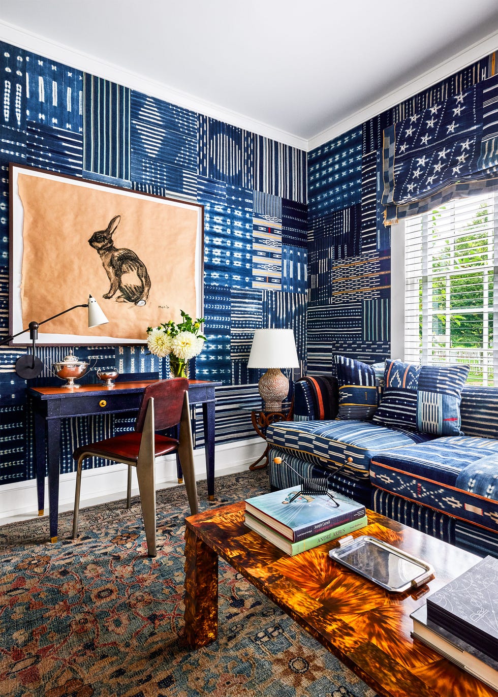 a den has walls and a sofa covered in quilted indigo textiles, window with blue shades and white stars, small orange desk with blue legs, flame colored cocktail table, persian rug, artwork of a rabbit