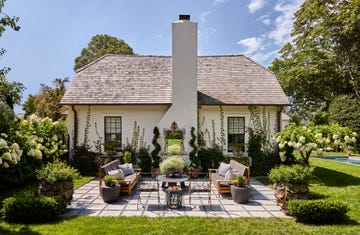 a cottage painted a light blush pink, chimney with a decorative mirror hanging on it, terrace with wood framed sofas with cushions, wrought iron side chairs and table, concrete tiles, flowering plants, grass lawn