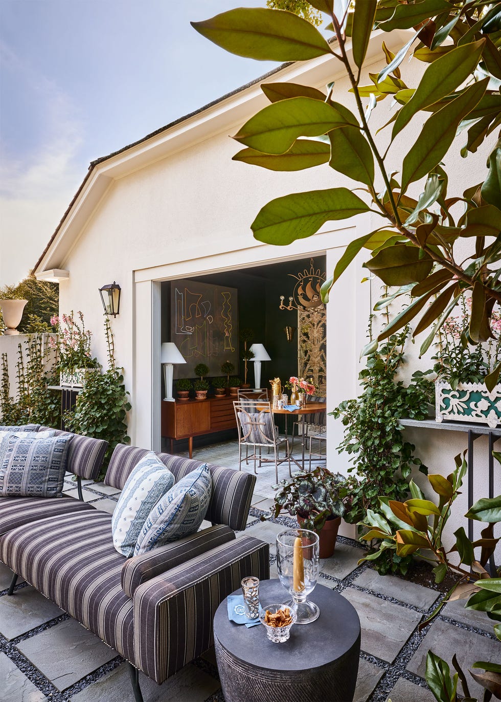 exterior of house opens to a terrace with square cement tiles set on gray pebbles, striped sofa with accent pillows, drumlike side table, flowering plants, inside house is a console and small dining table