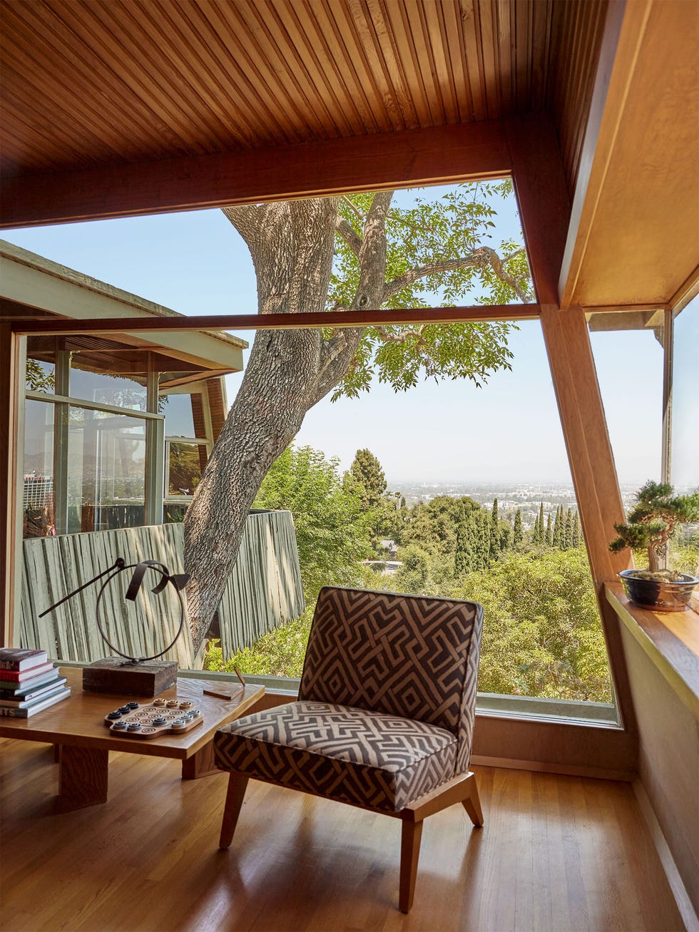 a brown patterned chair and a table in a room with a large glass window behind it and trees in view