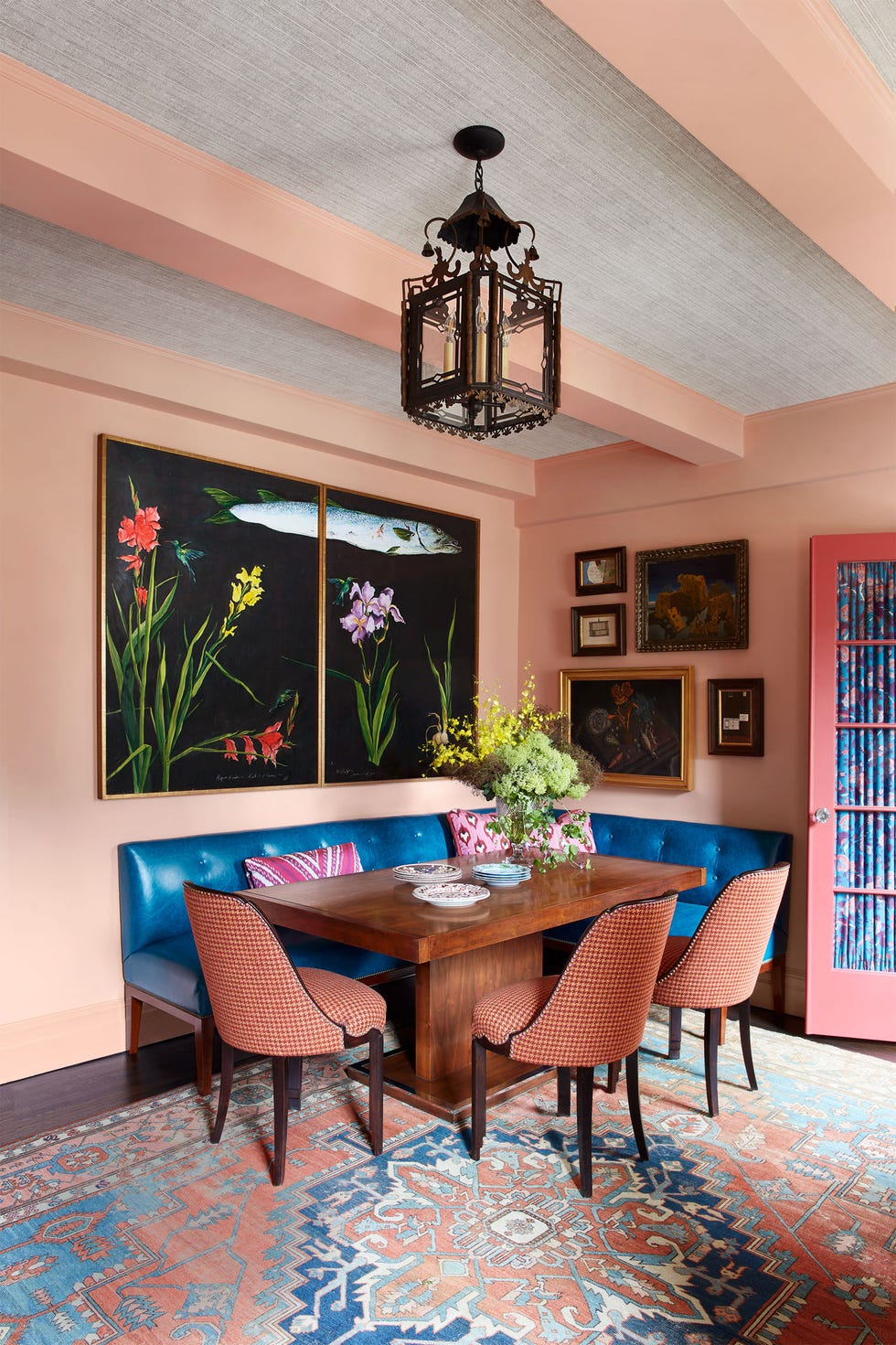 a light pink dining room with built-in blue leather bench, wooden table and three chairs in a spice-colored fabric, large diptych of flowers and fish, small framed art, antique metal pendants, oriental rug