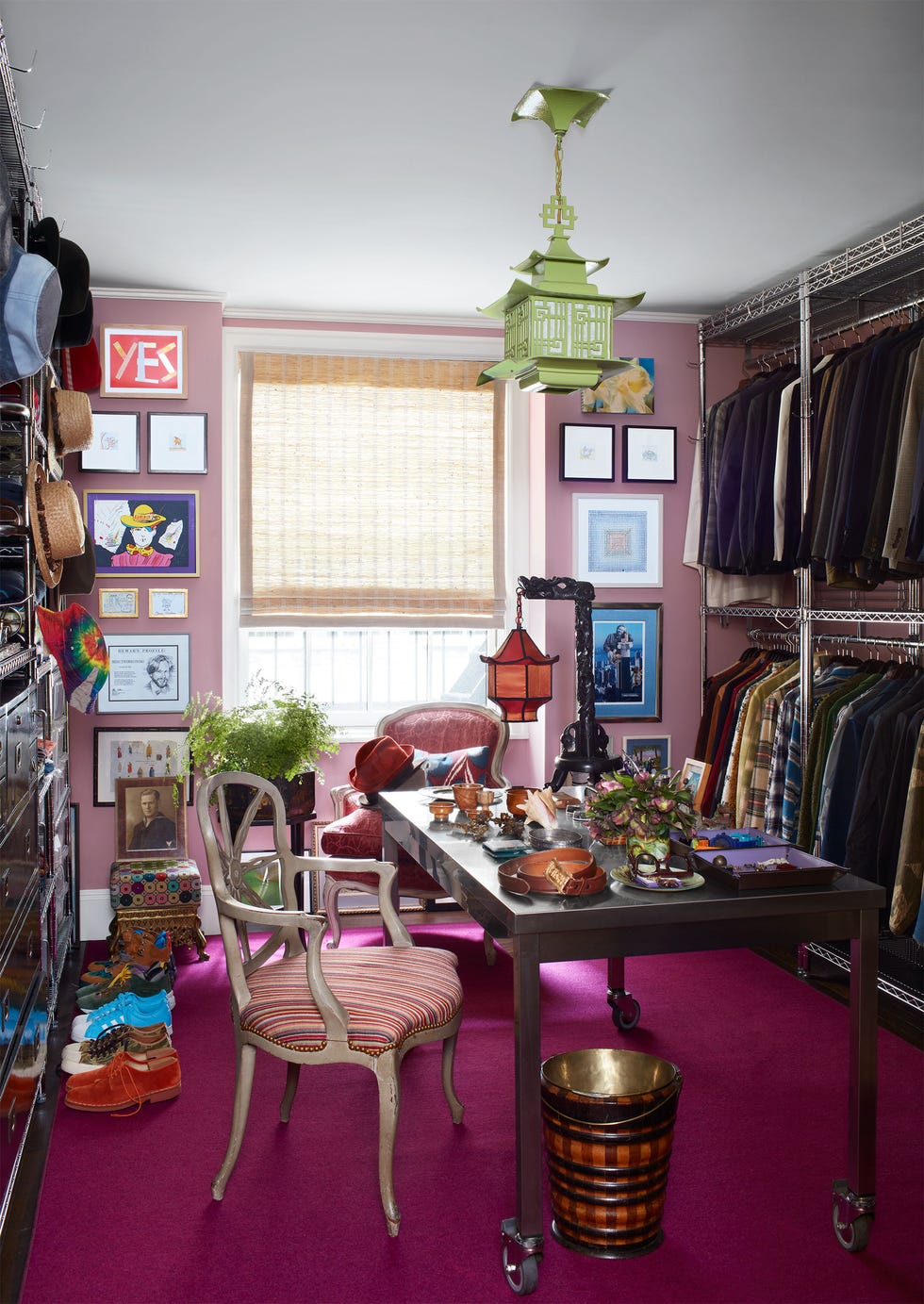 Dressing room with steel work table and antique chair, Chinese pendant light above, chair by the window with bamboo shade, magenta rug, pair of sneakers on the floor next to storage drawers, shirts hanging on rods