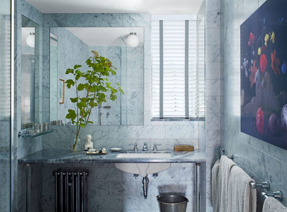 a bathroom with light marble walls and countertop, white sink with silver fittings, exposed sink and pipe and radiator under the countertop, mirror, globe lamp, small window with blind, towel rail, large floral artwork