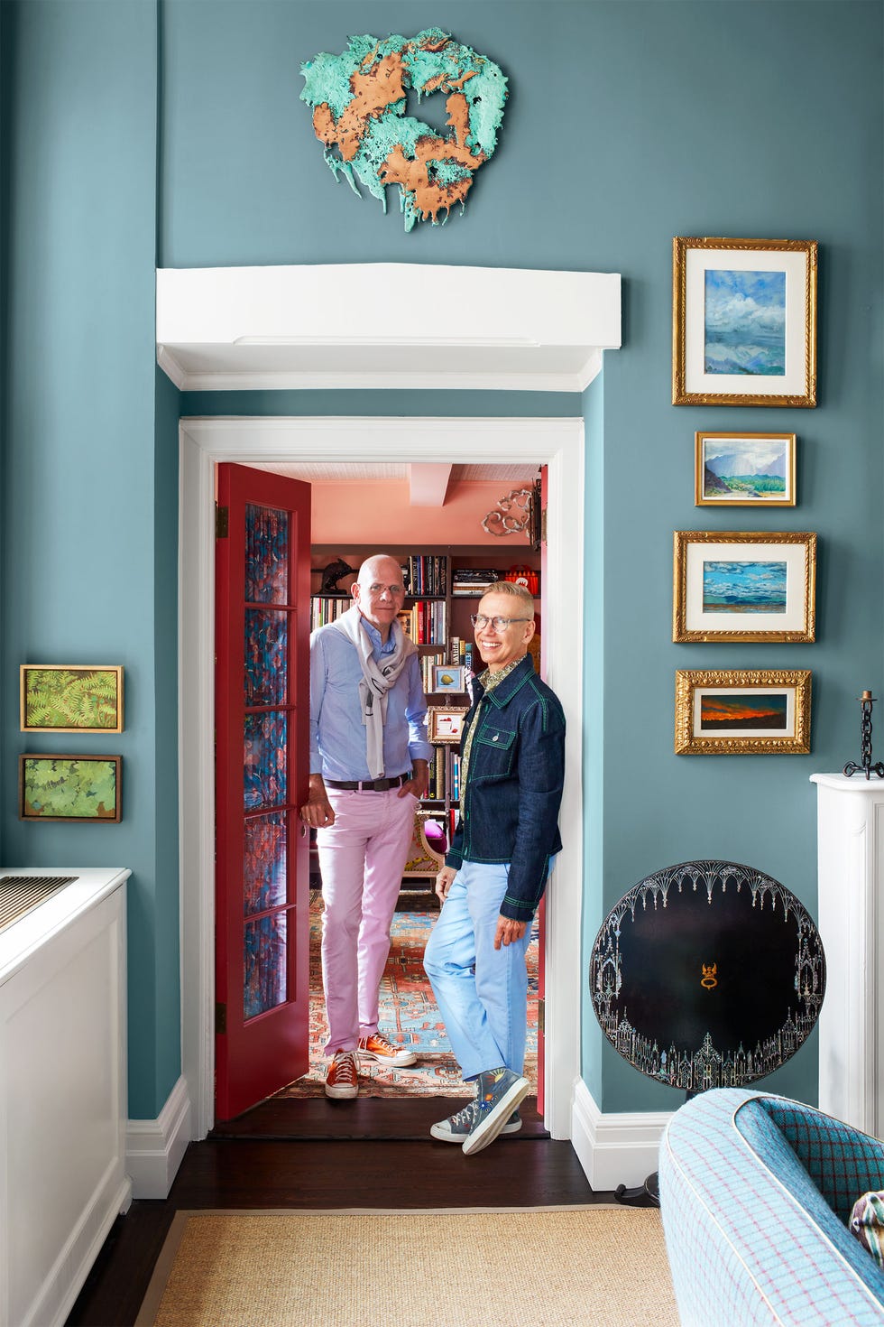 Two men stand in a doorway, one wearing a blue shirt, pink pants and red sneakers, the other wearing a denim jacket, light blue pants and blue sneakers. Above the doorway is a turquoise and copper-colored artwork.